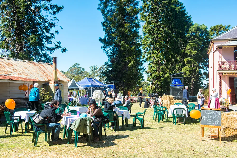 Outdoor farm event with families seated at green plastic chairs and tables on grass, hay bales, historic buildings, and blue tents under tall shady trees on a sunny day.
