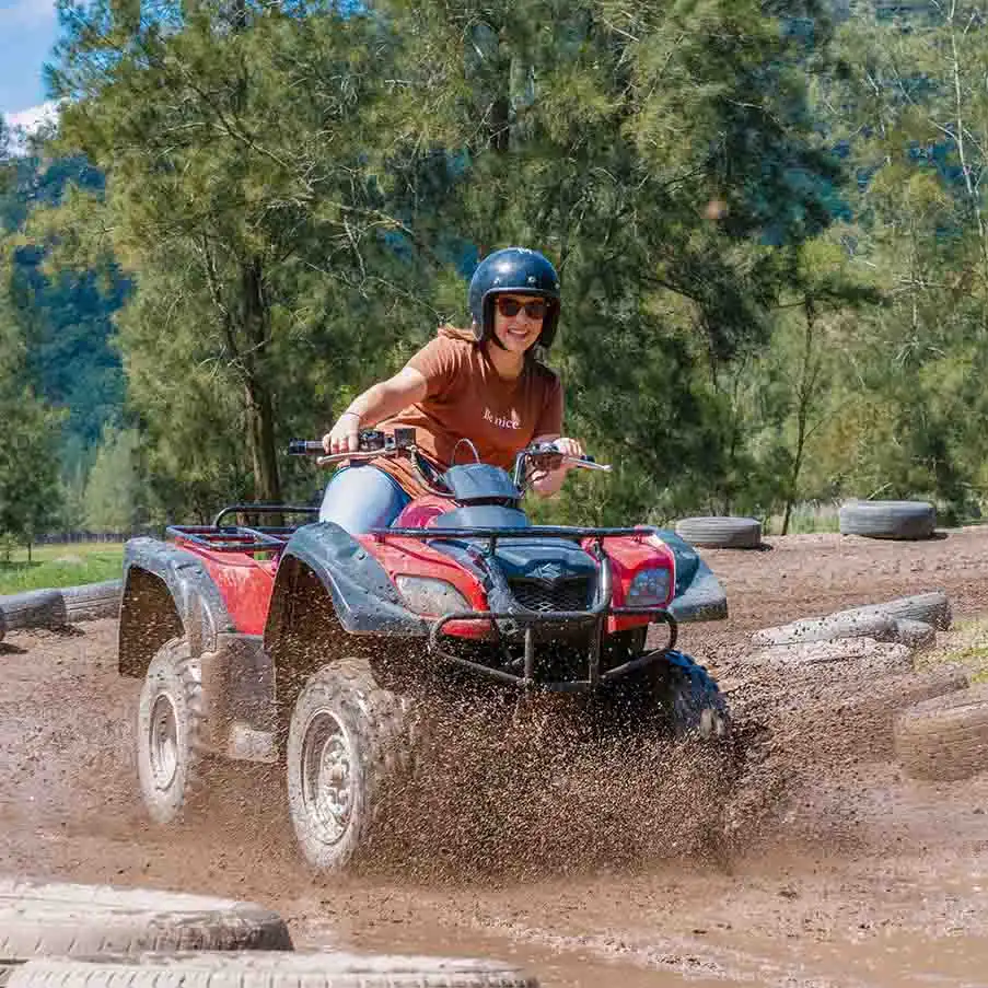 Woman in helmet riding red ATV through muddy terrain at outdoor adventure park, kicking up dirt with trees in background