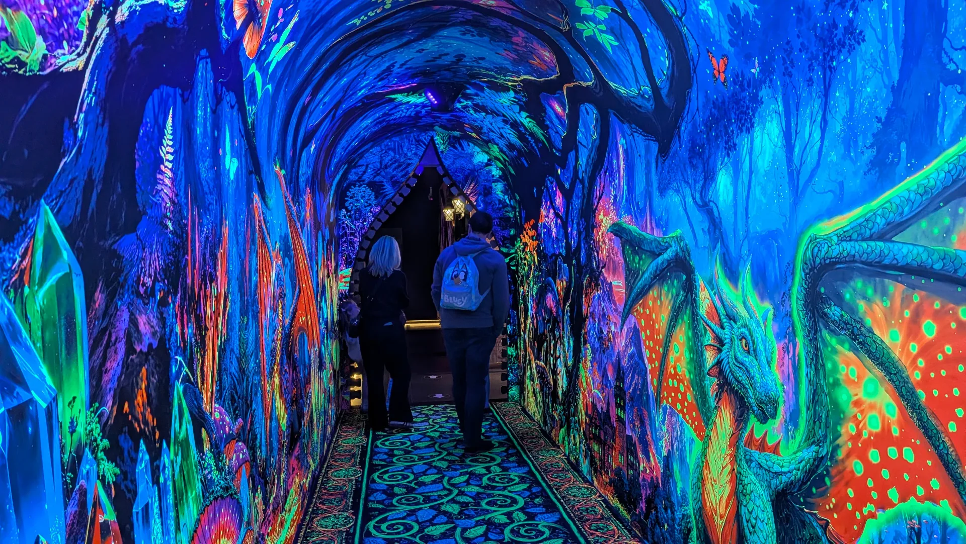 Vibrant fluorescent tunnel walkway with psychedelic blue, green and orange marine-themed artwork covering walls and ceiling, creating an immersive underwater experience for visitors.
