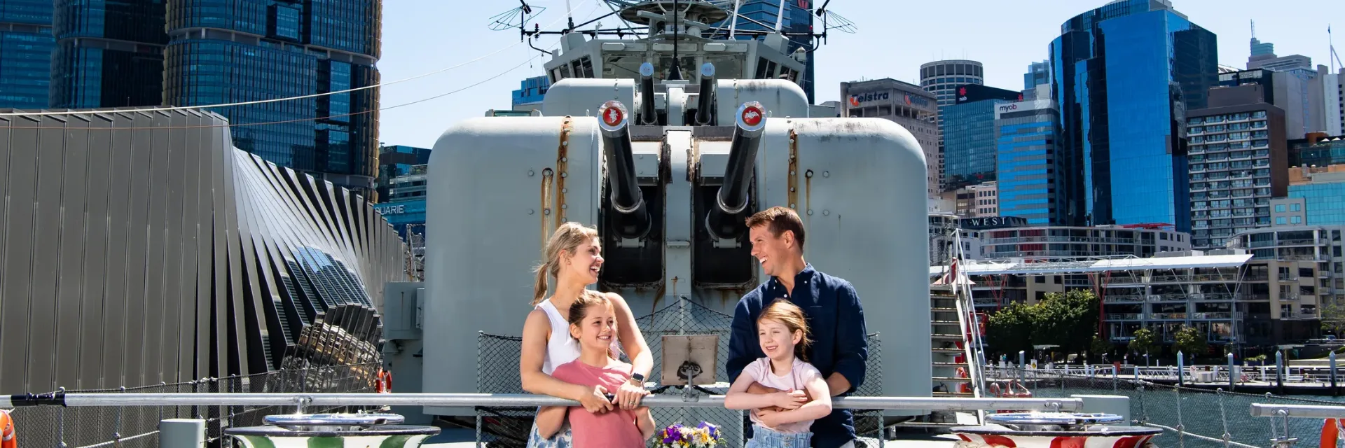 Family of four exploring the deck of a historic naval warship with city skyline backdrop at Australian National Maritime Museum
