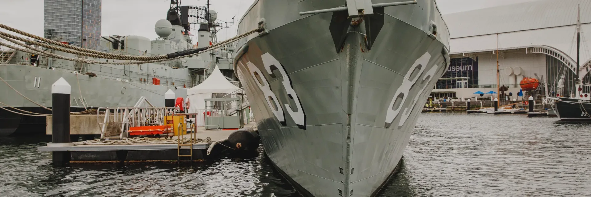 Gray naval warship with hull number 83 docked at Australian National Maritime Museum, featuring visitor walkways and white tent for educational activities