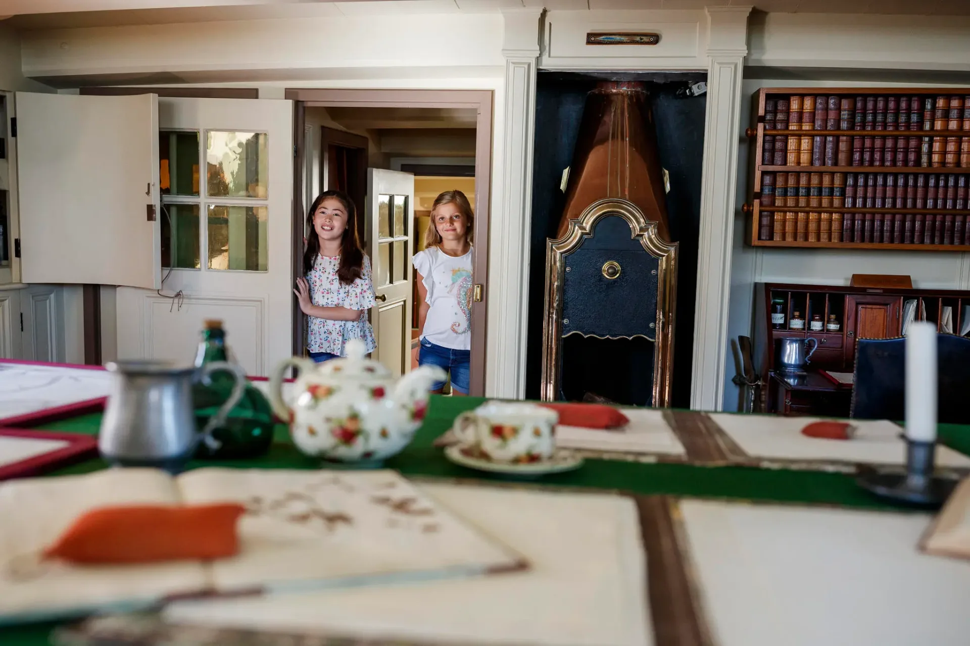 Two children exploring an interactive maritime exhibit featuring a large wooden ship's wheel display and nautical artifacts, with extensive bookshelves visible in the museum's learning space.