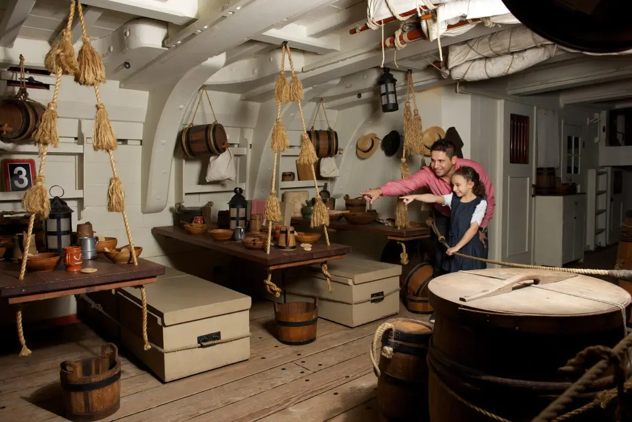 Father and child explore interactive ship galley exhibit with hanging rope storage, wooden barrels, and nautical artifacts at maritime museum