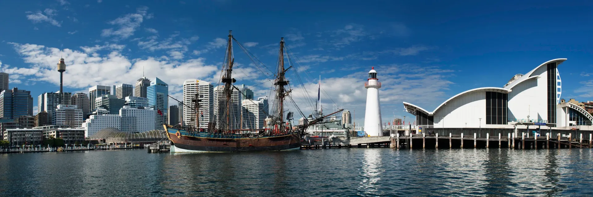 Historic tall ship docked at Darling Harbour with Sydney city skyline, lighthouse, and modern maritime museum buildings surrounding the waterfront