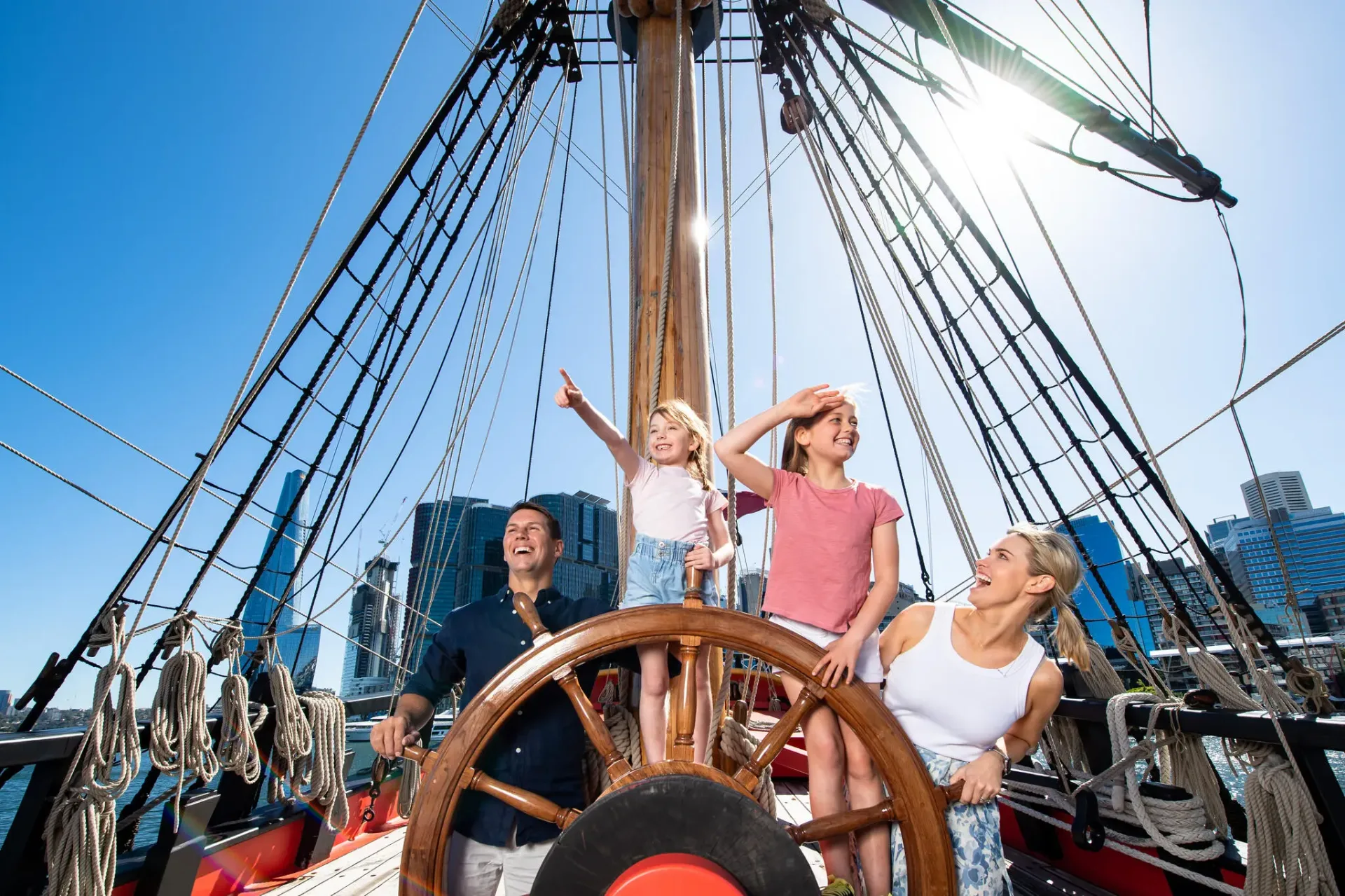 Family of four explores the deck of a historic tall ship at Australian National Maritime Museum, with children at the ship's wheel and Sydney harbor skyline visible in background