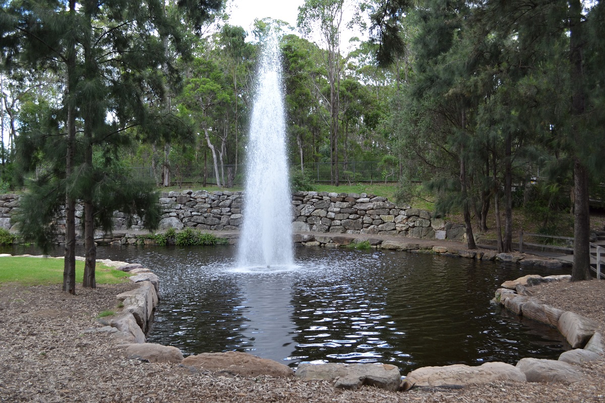 Fountain spraying upward in a rock-walled pond ringed with sandstone boulders and surrounded by tall native trees.