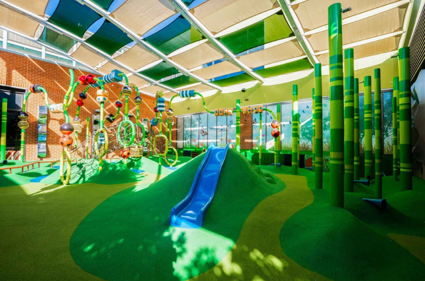 The Backyard playground at Rouse Hill Town Centre — an open-air fenced play space with a blue slide built into a green soft-fall mound, climbing rings and musical play columns, near the Coles quad.