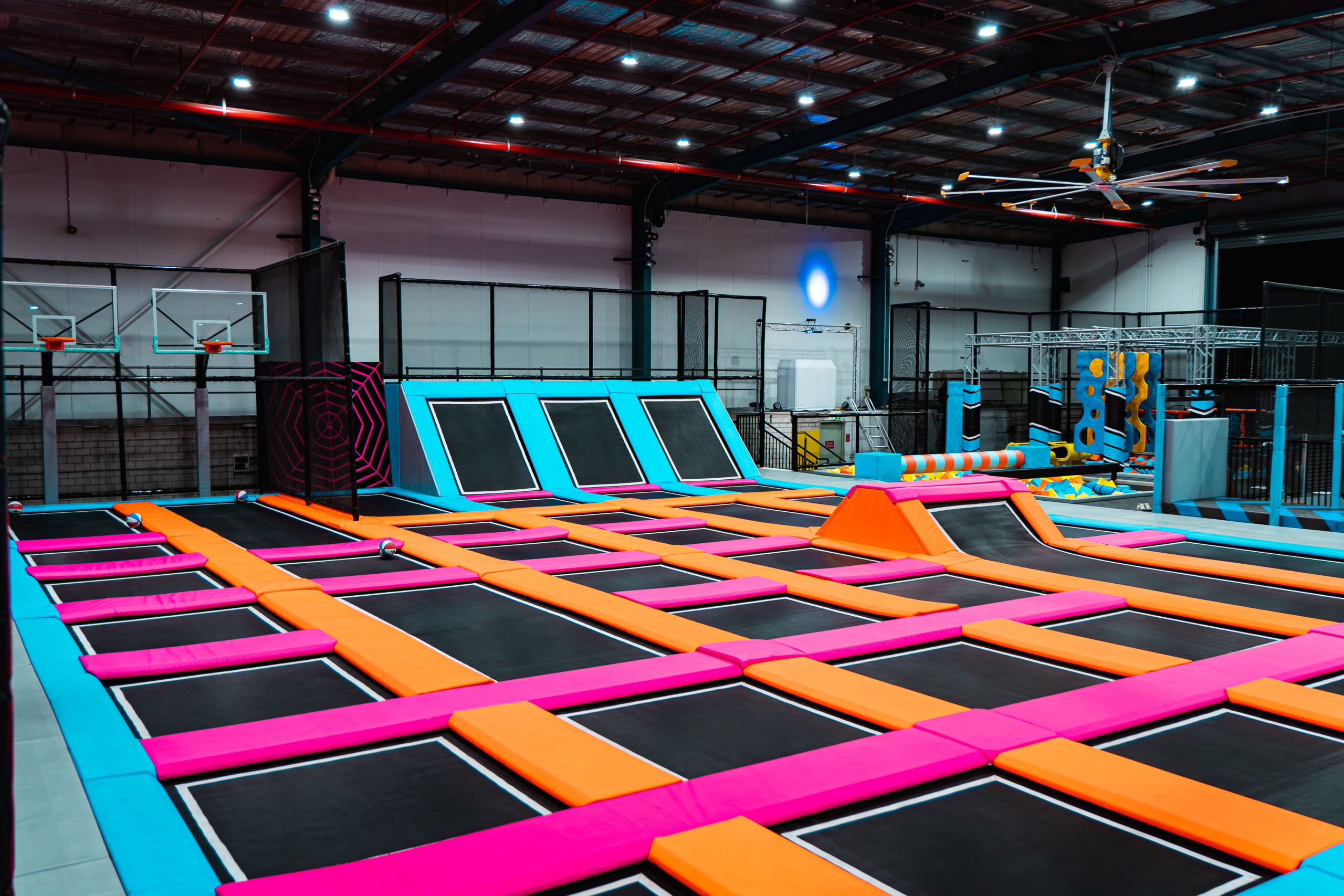 Wall-to-wall indoor trampoline arena with pink, orange and blue padded trampolines, angled wall-runs, a basketball dunk hoop and an angled foam jump area