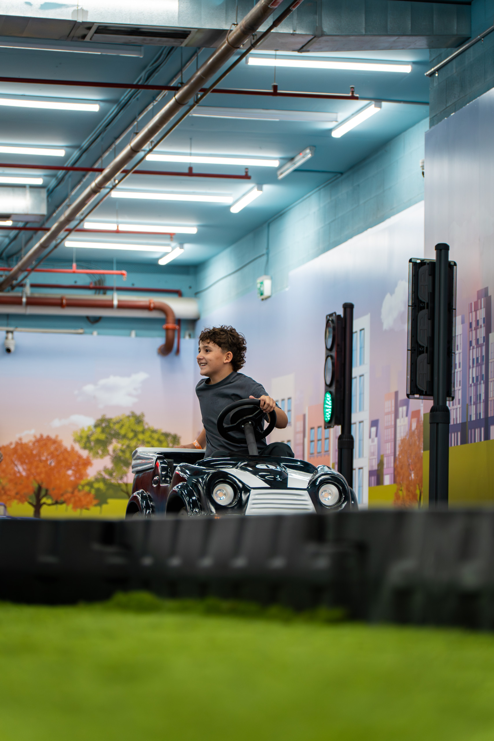 A child driving a small electric ride-on car along the indoor City Drive track with a painted streetscape mural and traffic lights
