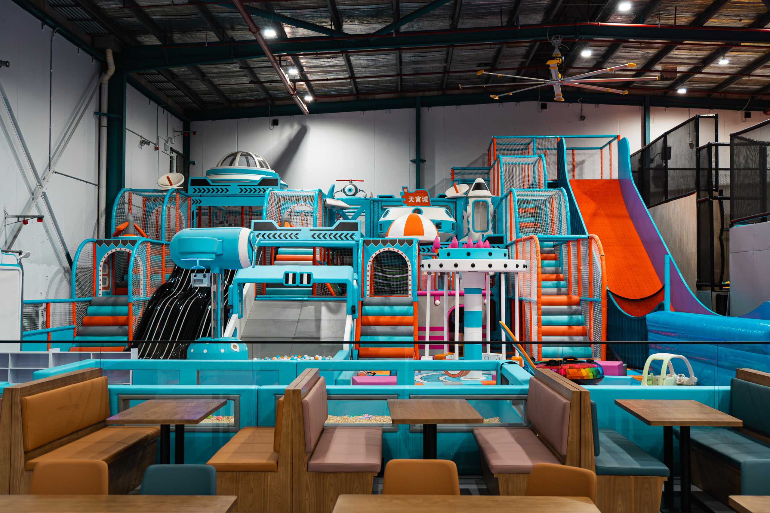 Towering blue and orange themed soft play structure with slides, viewed across booth-style cafe seating tables in the foreground