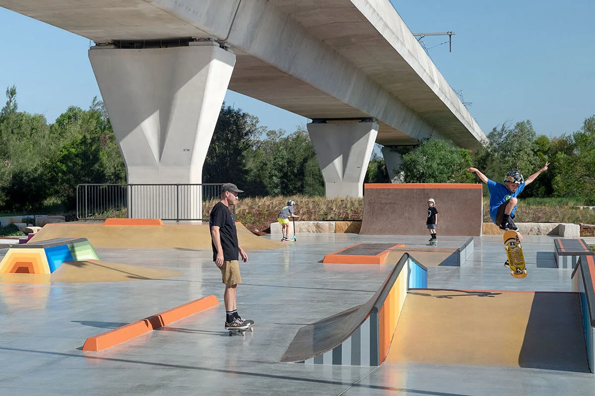Modern concrete skate park with colorful ramps and rails beneath a highway bridge, featuring skaters performing tricks while others practice on various obstacles.