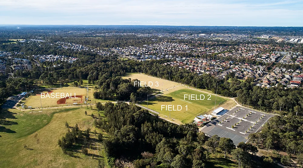 Aerial view of Caddies Creek Reserve showing multiple sports fields including baseball diamond, three numbered grass fields, parking area, and surrounding residential neighborhoods with tree coverage.