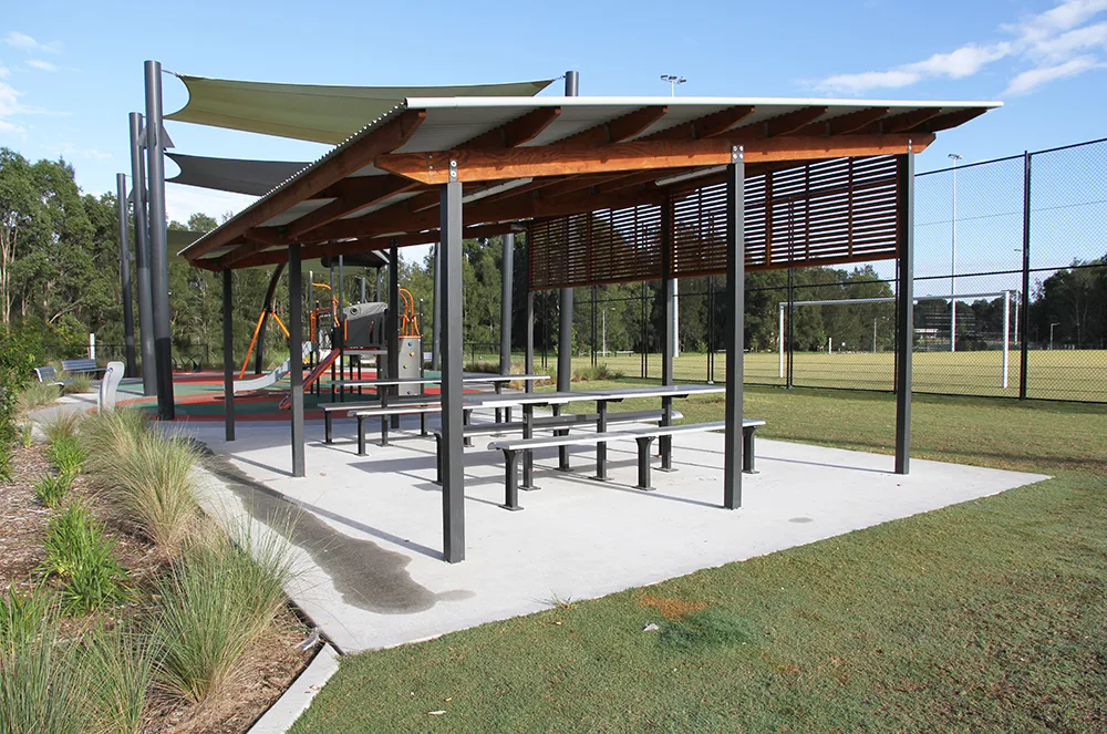 Modern covered shelter with picnic tables on concrete pad, shade sails, and playground equipment visible in background at park reserve