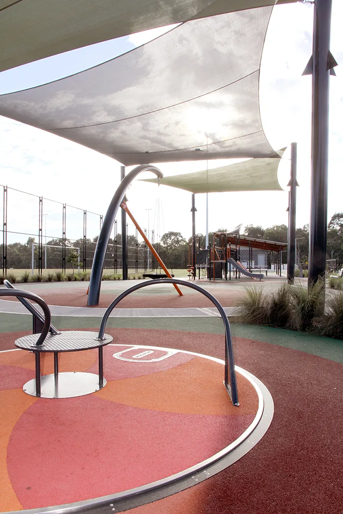 Modern playground with curved metal play equipment on colorful rubber surfacing under large shade sails, with additional playground structures visible in background