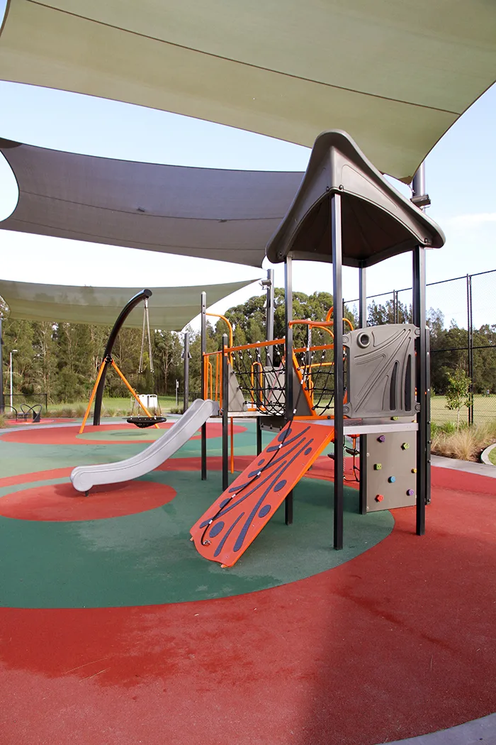 Modern playground with curved slide, climbing wall, and rope features under large shade sails on colorful rubber surfacing at Caddies Creek Reserve