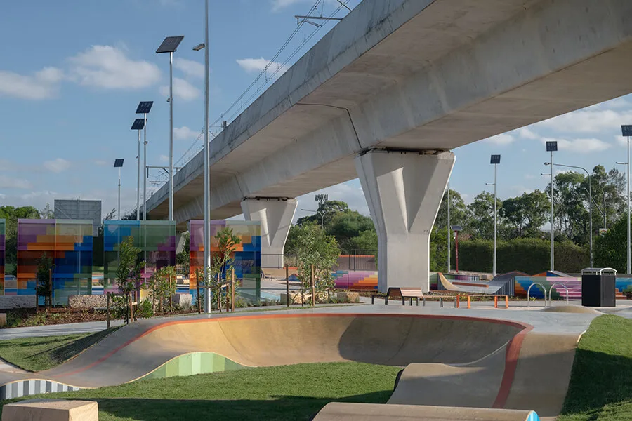 Modern concrete skate park with curved bowls and ramps beneath an elevated train bridge, featuring colorful street art murals and solar-powered lighting poles in a landscaped setting.