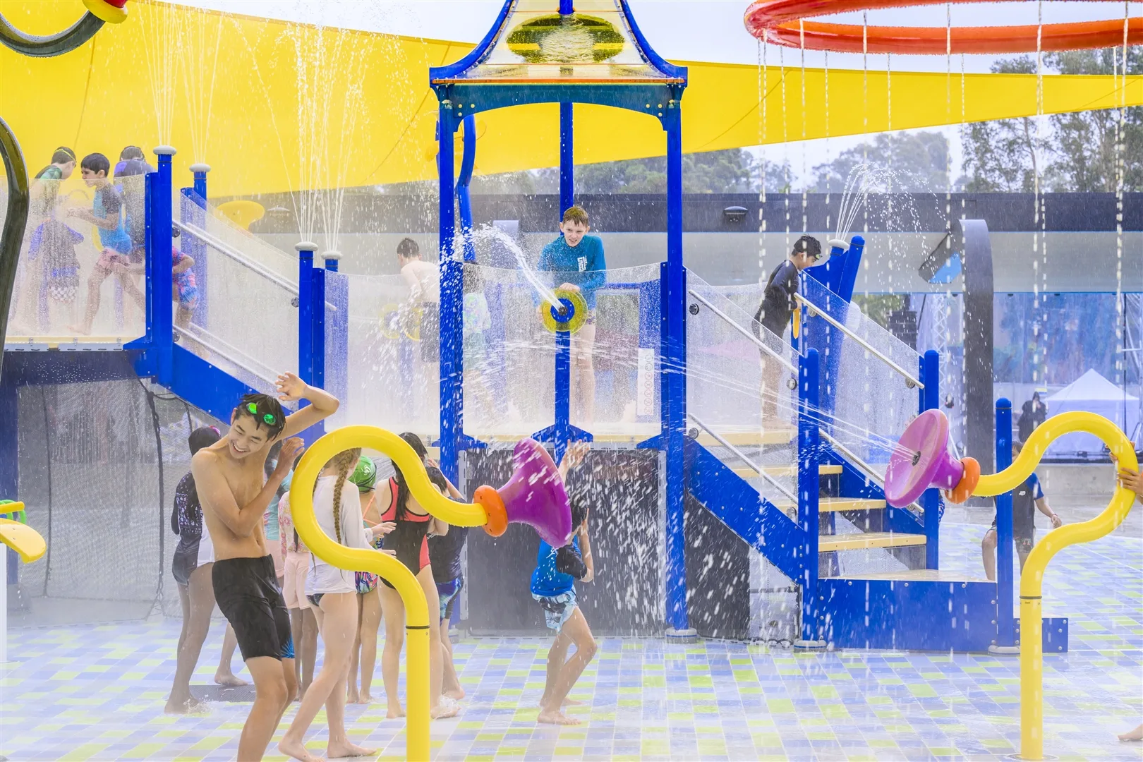 Children playing in a colorful water playground with blue climbing structure, yellow water features, and multiple spray fountains at an indoor aquatic center