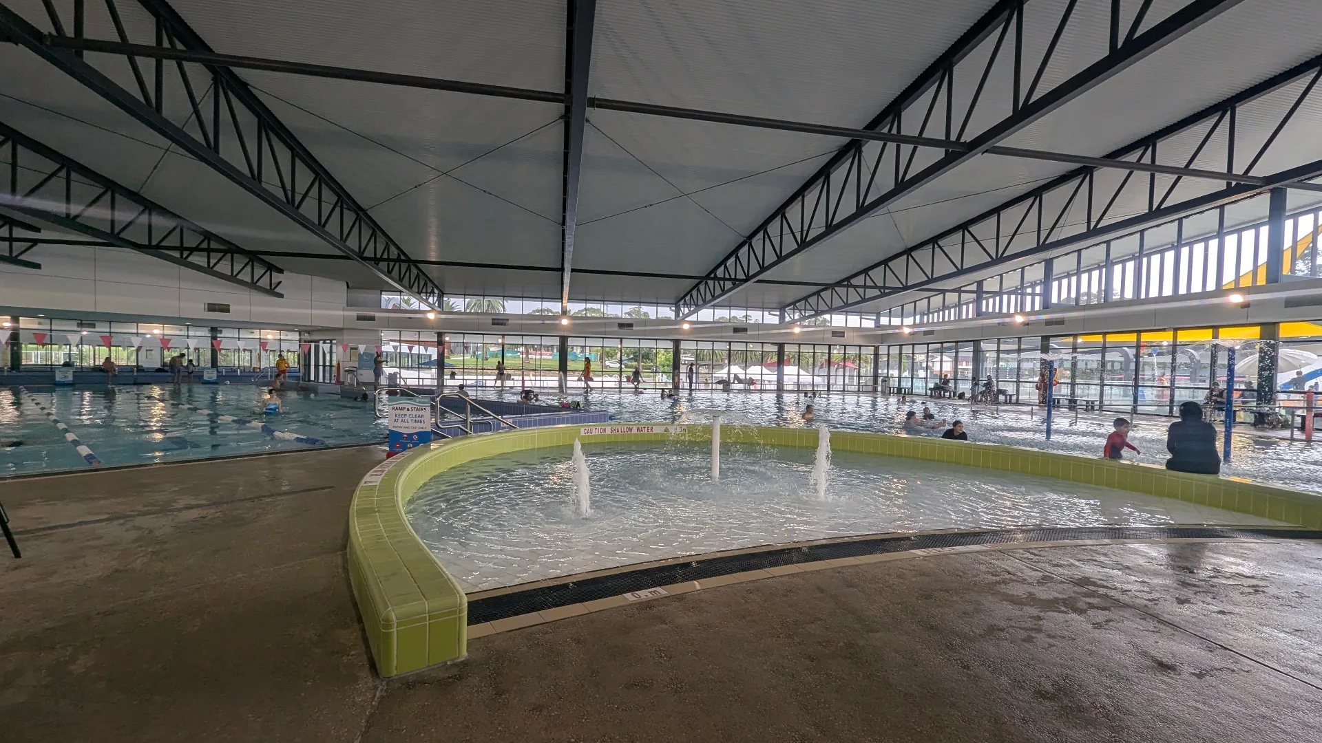 Indoor aquatic center with circular shallow splash pad featuring water fountains, lap pool with lane ropes, and covered seating areas under steel beam ceiling