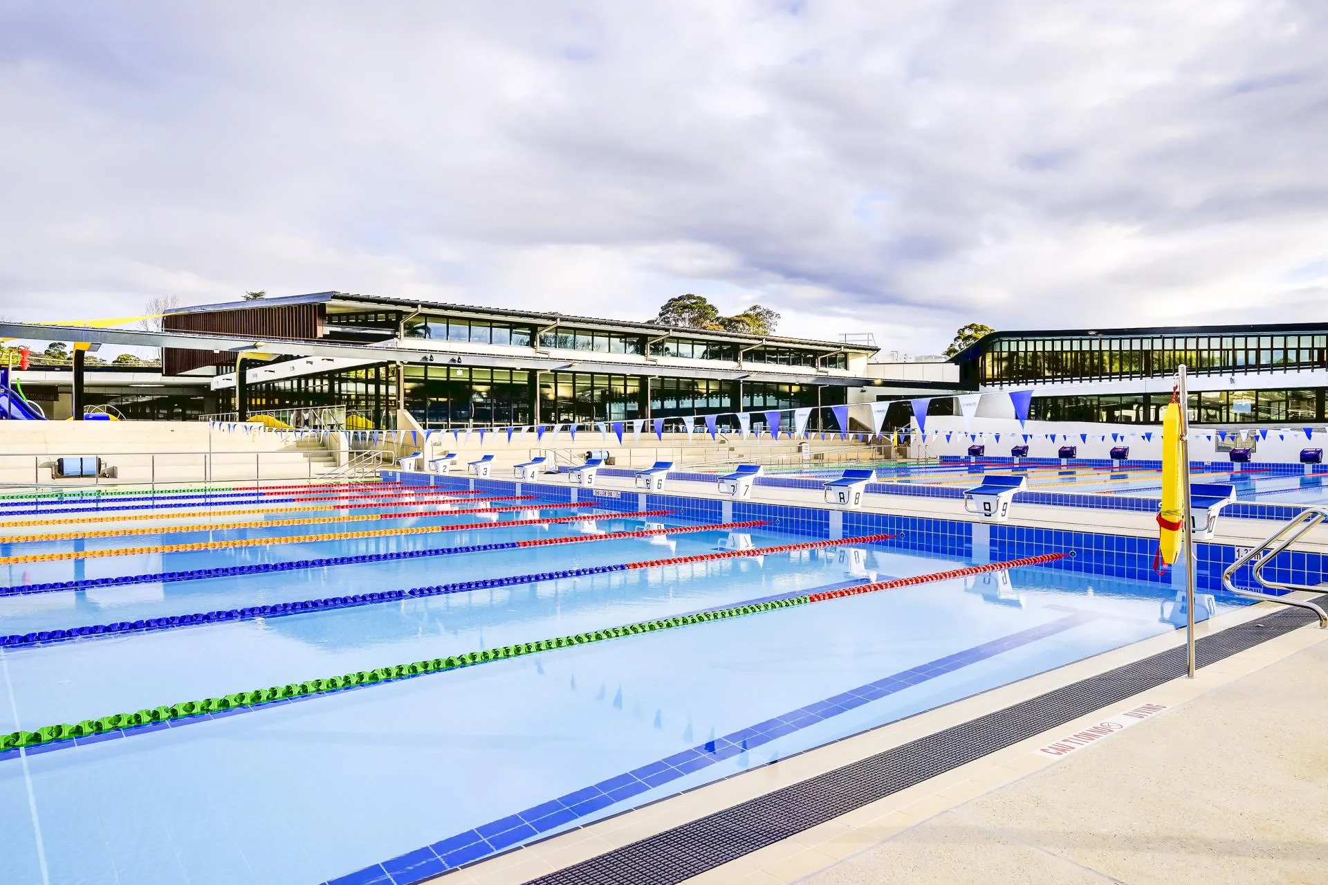 Modern aquatic center with Olympic-sized swimming pool featuring multiple lanes marked by colorful rope dividers, stadium seating, and contemporary glass buildings with covered spectator areas.