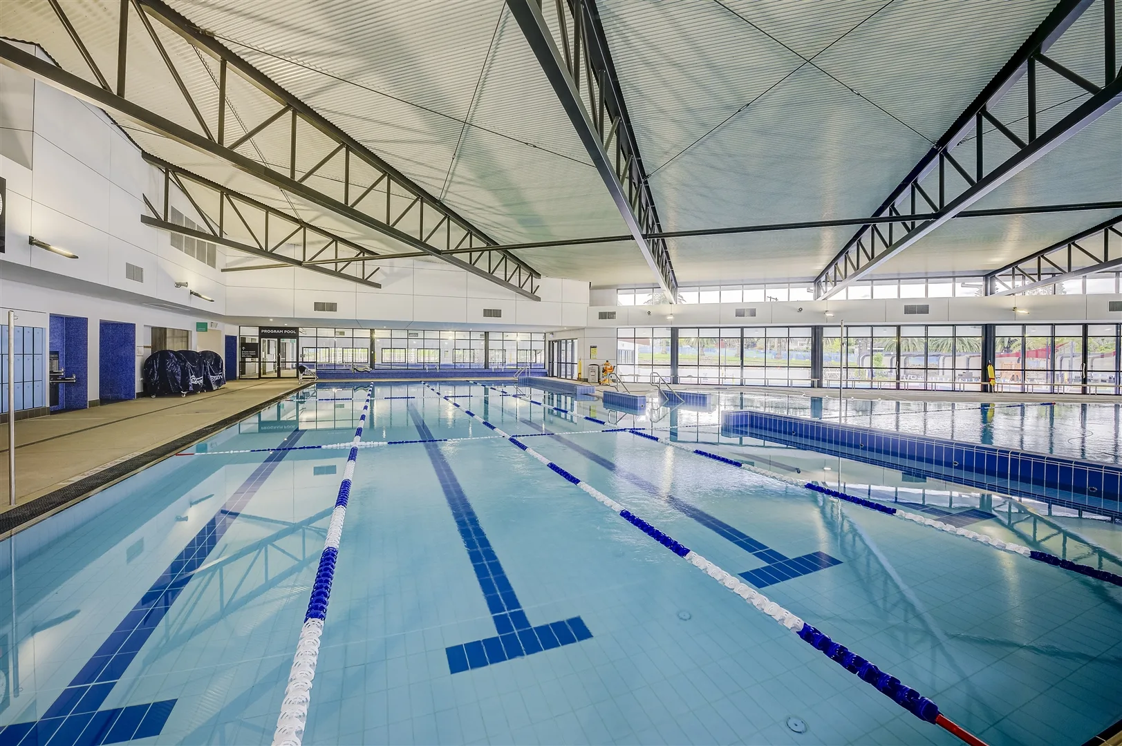 Indoor swimming pool with multiple lap lanes marked by blue and white lane ropes, featuring high ceilings with natural light from large windows and modern architectural steel beams.