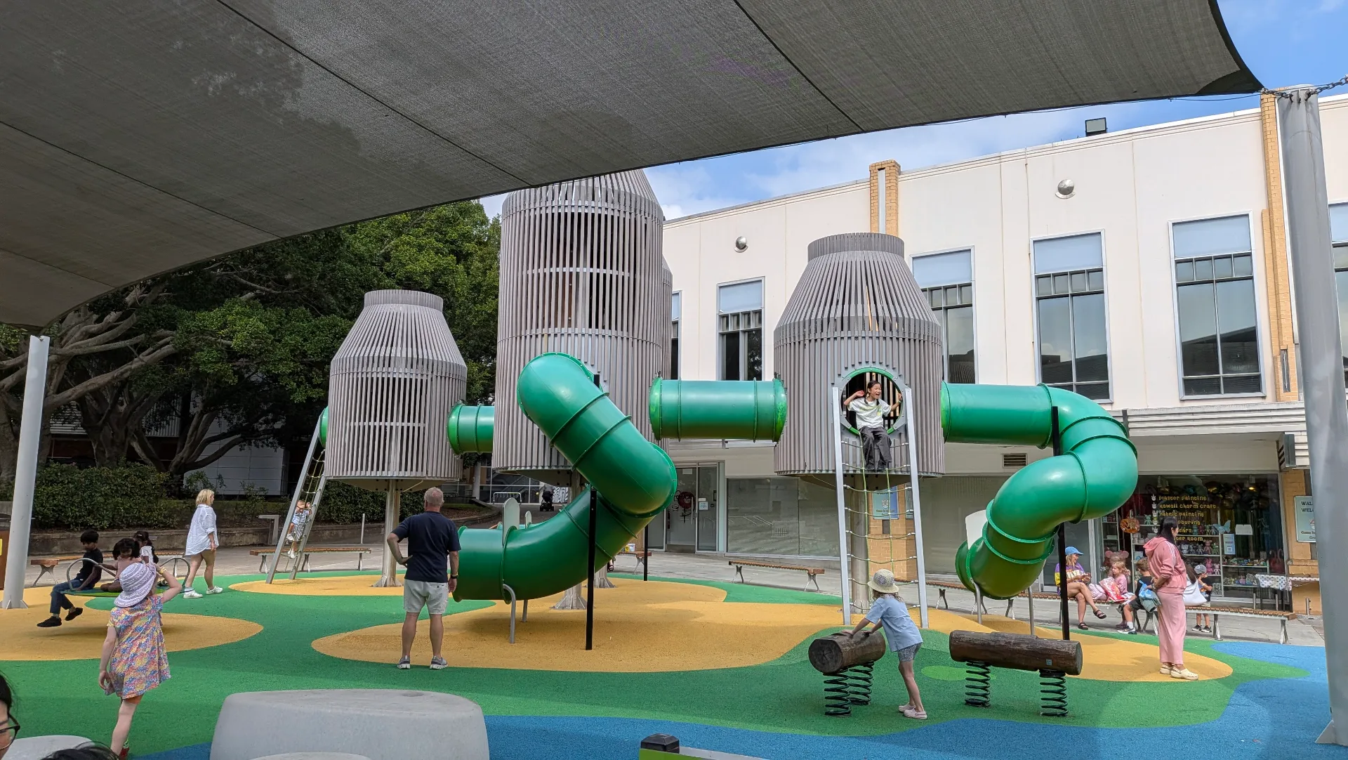 Modern playground with large green tube slides winding between tall wooden tower structures, colorful rubber safety surfacing, and families playing under shade sails.