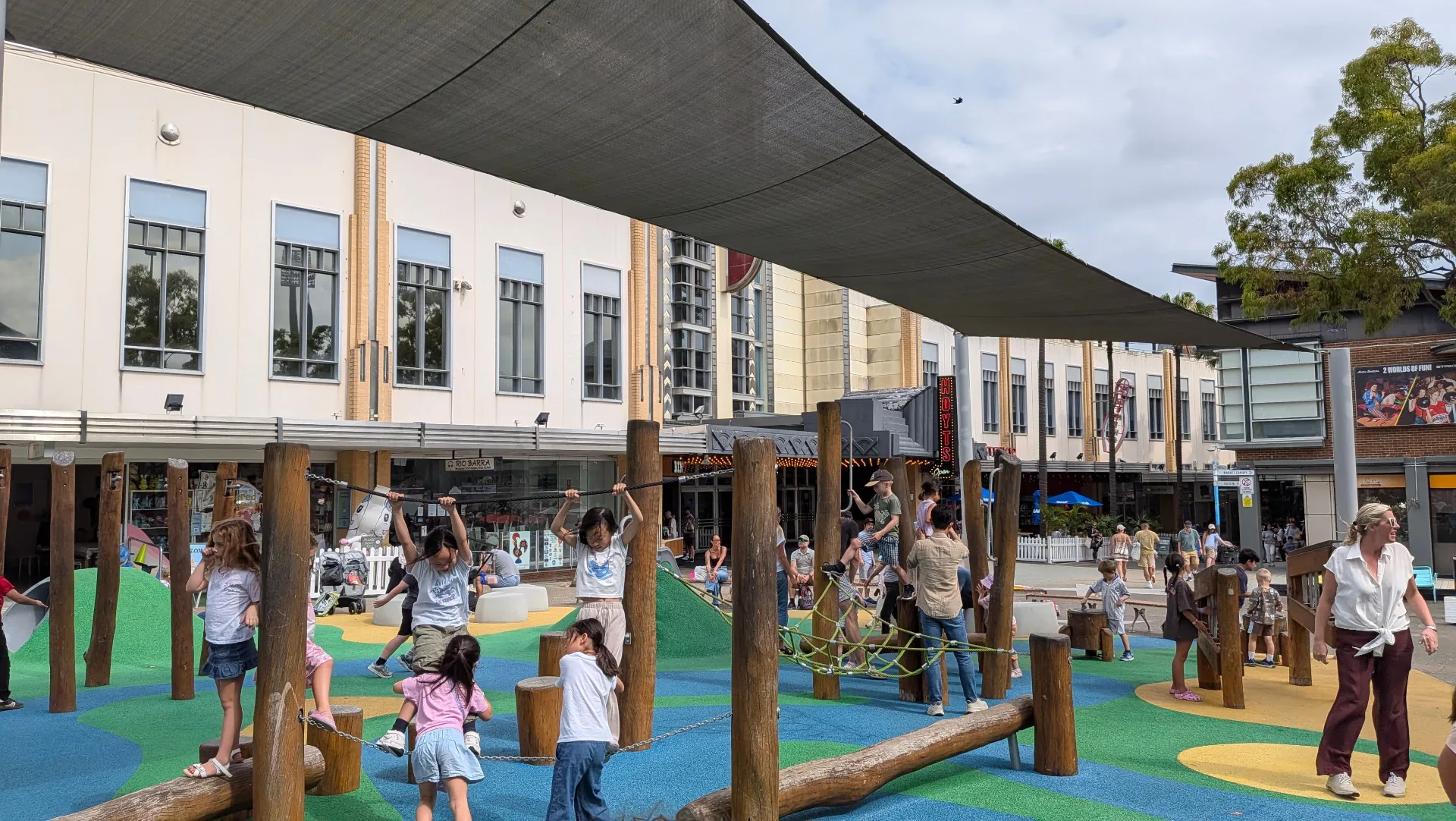 Children playing on a natural timber playground with rope bridges and climbing posts under shade sails, set in a town square with shops visible in the background.