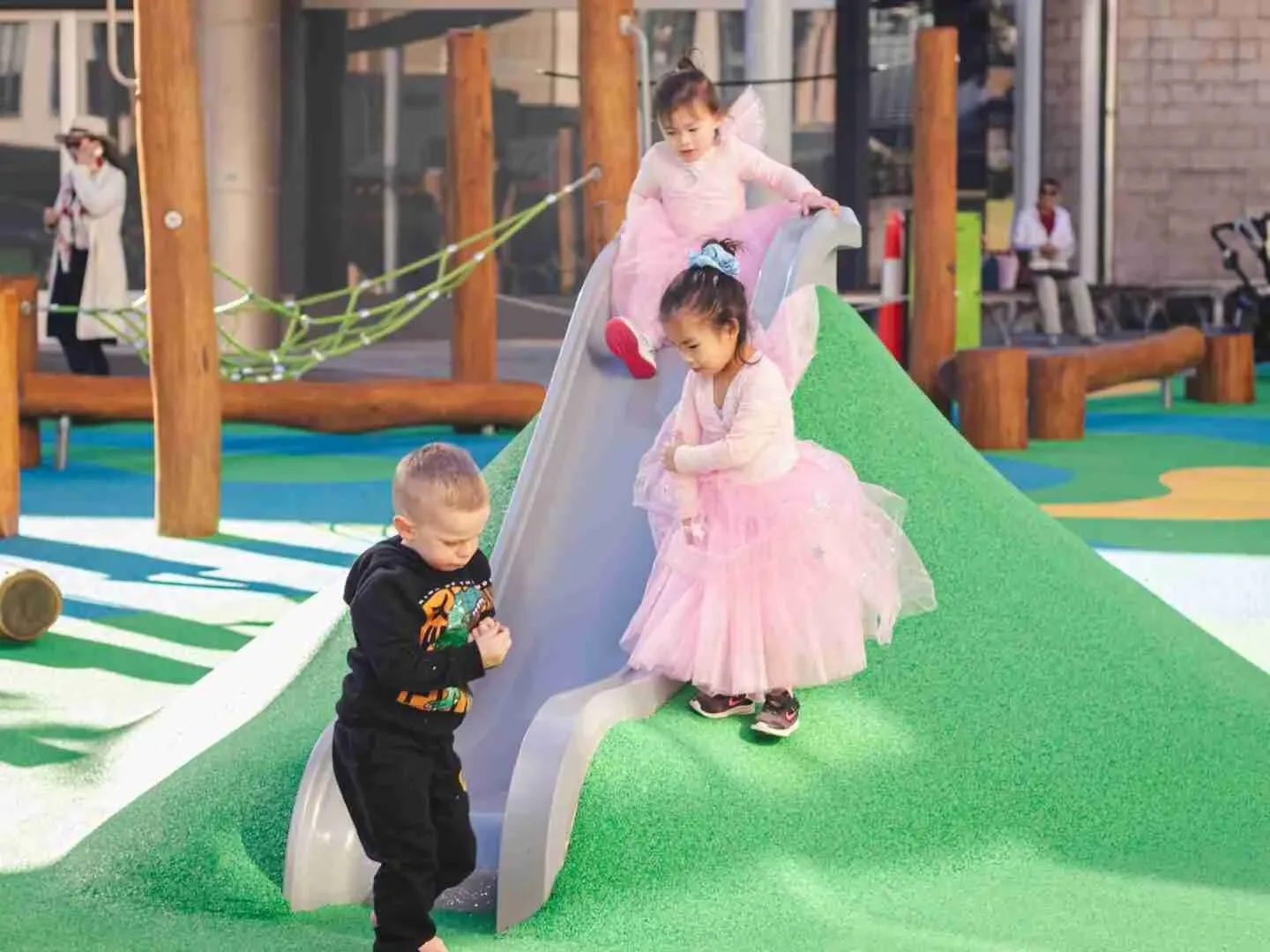 Children playing on a colorful playground with gray slide and green artificial turf surface, including a boy in dark clothing and girls in pink dresses at modern play structure.