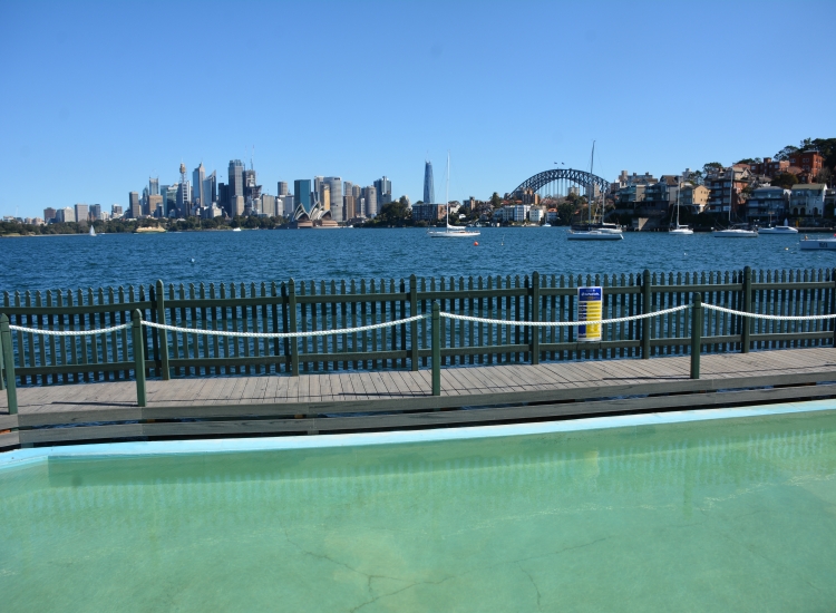 MacCallum Pool's seawater fills the foreground beside a timber deck and picket safety fence, looking across Sydney Harbour to the city skyline, Opera House and Harbour Bridge under a clear sky.