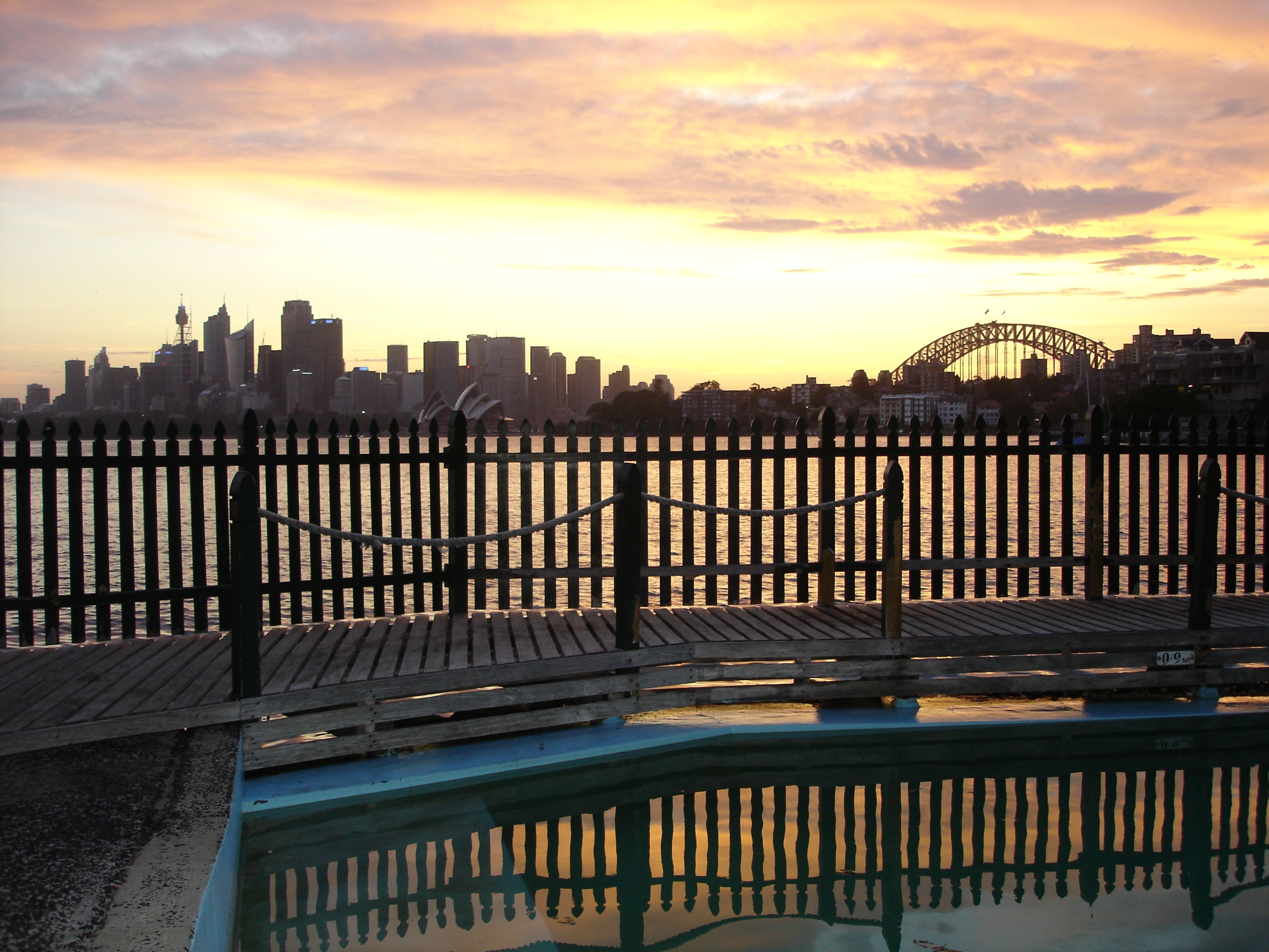 Sunset over Sydney Harbour seen from the MacCallum Pool deck, with the pool water and a silhouetted safety fence in the foreground and the city skyline and Harbour Bridge on the horizon.