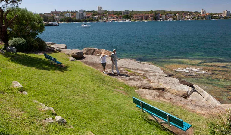 Waterfront park with grassy area, bench seating, and rocky shoreline where two people stand near calm harbor waters with city skyline visible across the bay