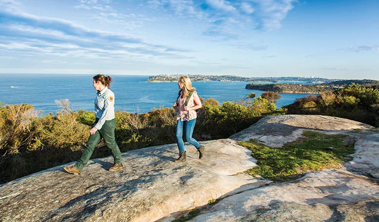Two women hiking along coastal rock formations with panoramic ocean views and distant headlands, featuring natural stone paths suitable for families exploring the scenic Spit to Manly Walk.