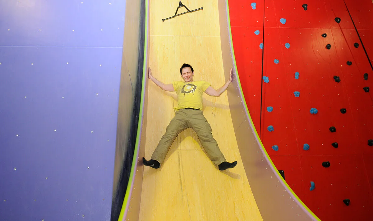 A man smiles while sliding down a large, steep yellow vertical drop slide between colorful climbing walls.