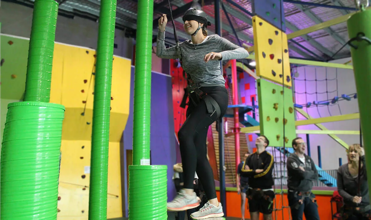 A woman in a harness and helmet balances across high green stepping pillars in an indoor ropes course.