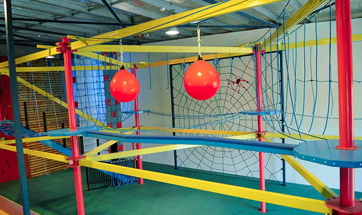 Indoor aerial adventure park featuring yellow beams, blue walking tracks, red support poles, and a spider web climbing net.