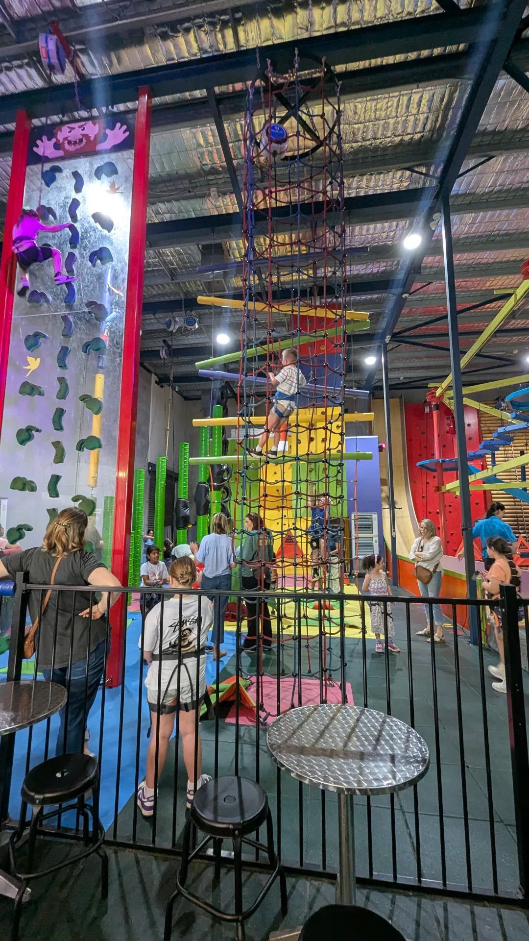 Vibrant indoor play center with children using a vertical net climb, various climbing walls, and a fenced-off cafe seating area.