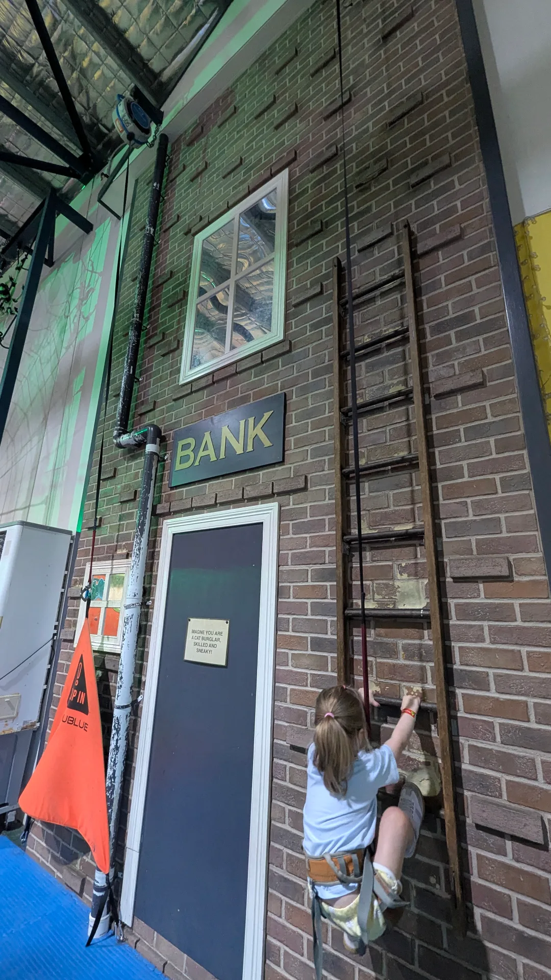 A young girl in a safety harness climbs a brick-themed "Bank" wall using a wooden ladder and wall grips.