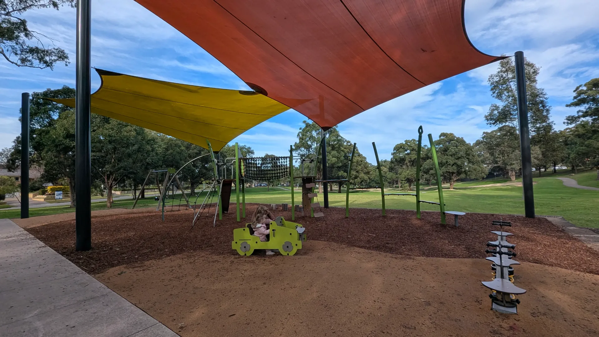 A wide view of the playground featuring climbing nets, a swing set, and a jeep rocker, all protected by large overhead shade sails.