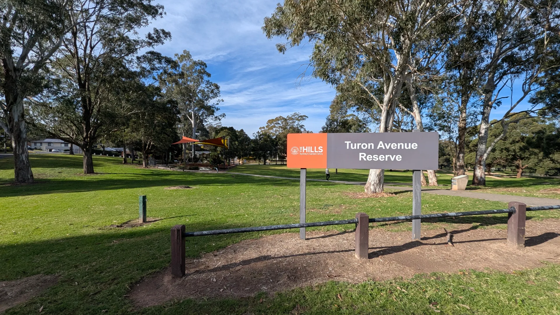 A grey and orange sign for "Turon Avenue Reserve" standing in a lush green park with tall trees and a shaded playground in the distance.