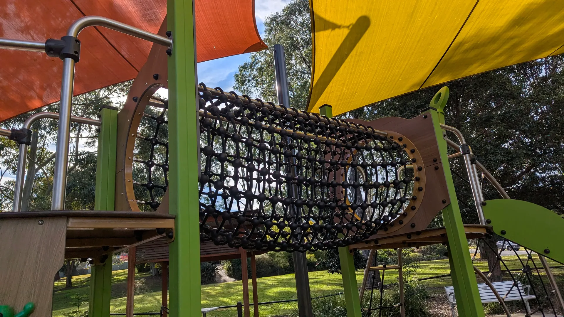 A black rope-tunnel climbing bridge connecting two elevated wooden platforms under bright orange and yellow shade sails.