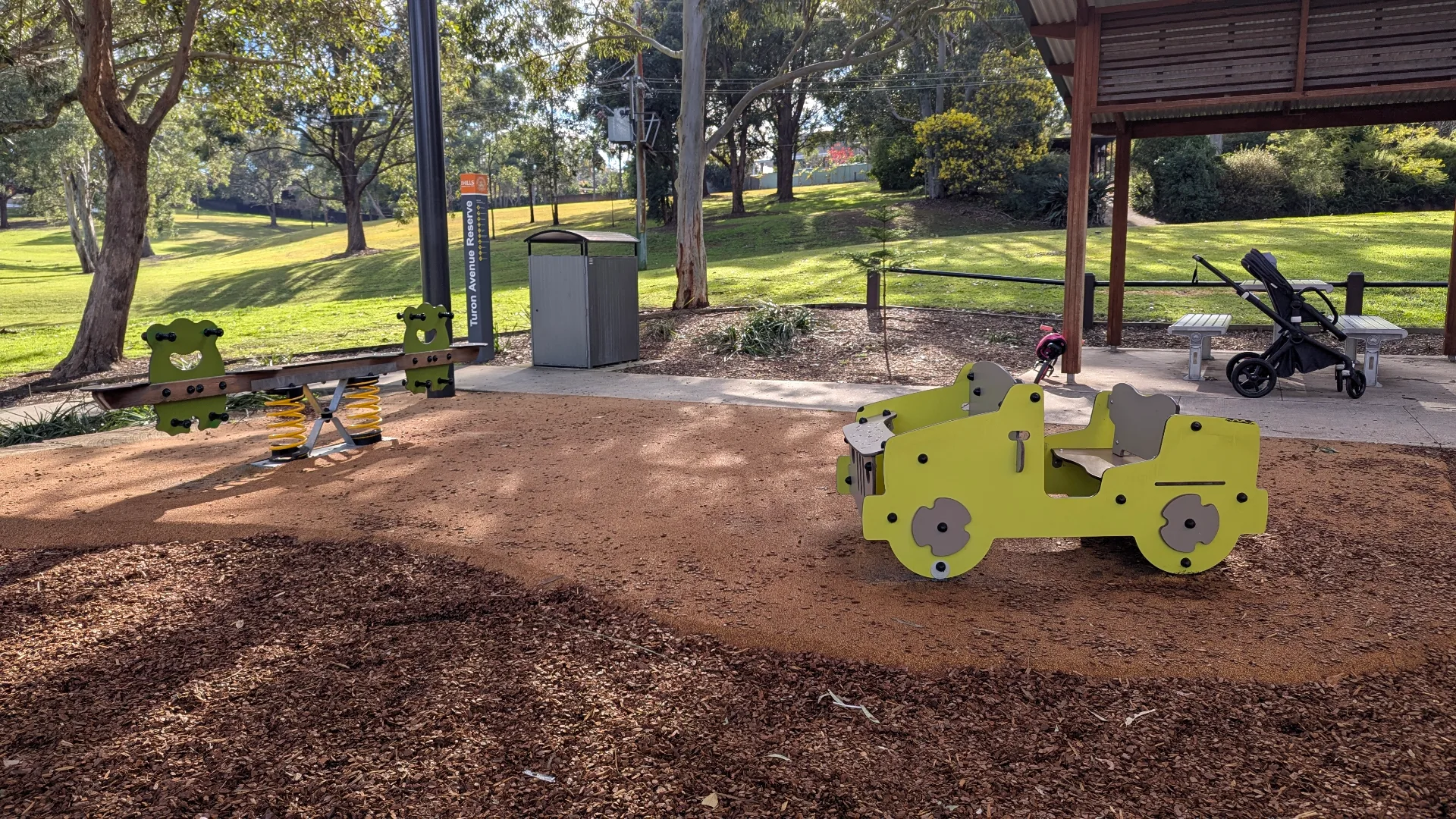A green spring-loaded seesaw and a jeep-themed toddler play structure on a mix of soft rubber and bark mulch flooring.