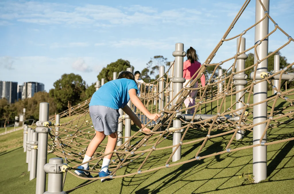 Children navigating a large, low-to-the-ground rope climbing net structure set over soft green artificial turf.