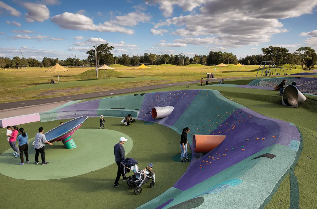 Families exploring a colorful, undulating play space with soft rubber flooring, built-in play tunnels, and a spinning disk.