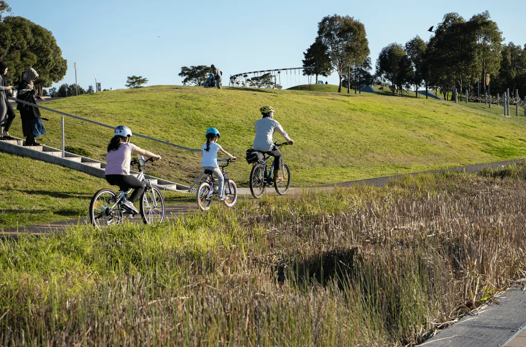 A family rides bicycles along a paved, stroller-friendly path winding through grassy hills and natural wetlands.