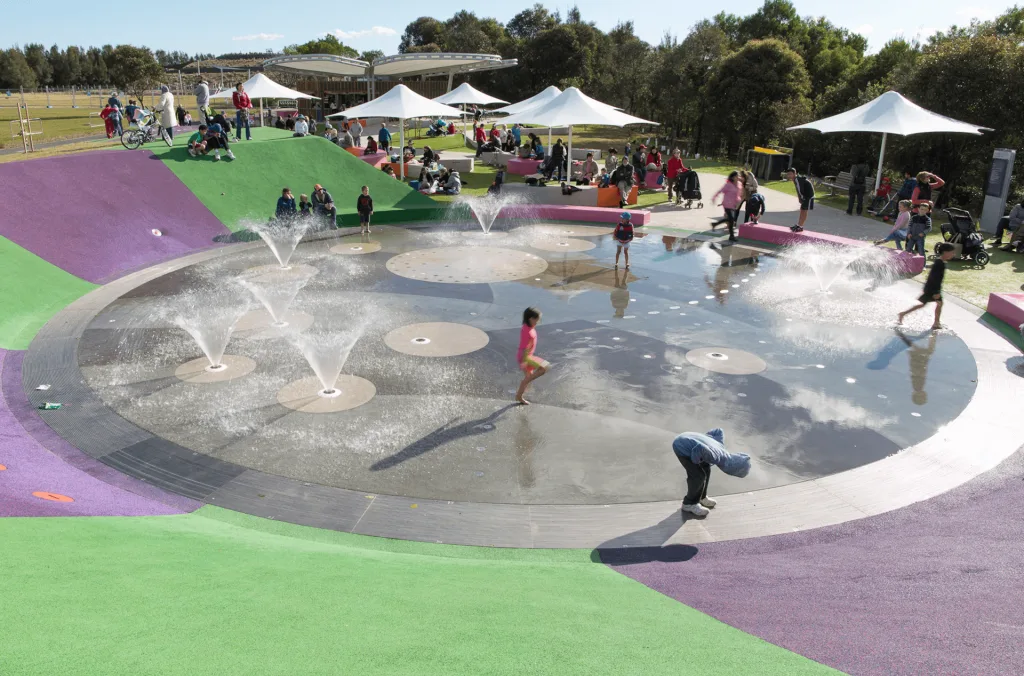 Kids playing in a vibrant circular splash pad with ground-level water jets, surrounded by rubber flooring and shade umbrellas.