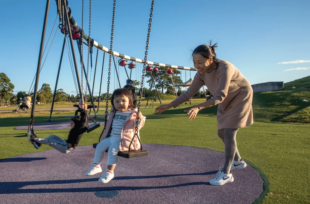 Smiling mother pushing a toddler on a bucket swing under a large multi-swing structure with soft rubber flooring.