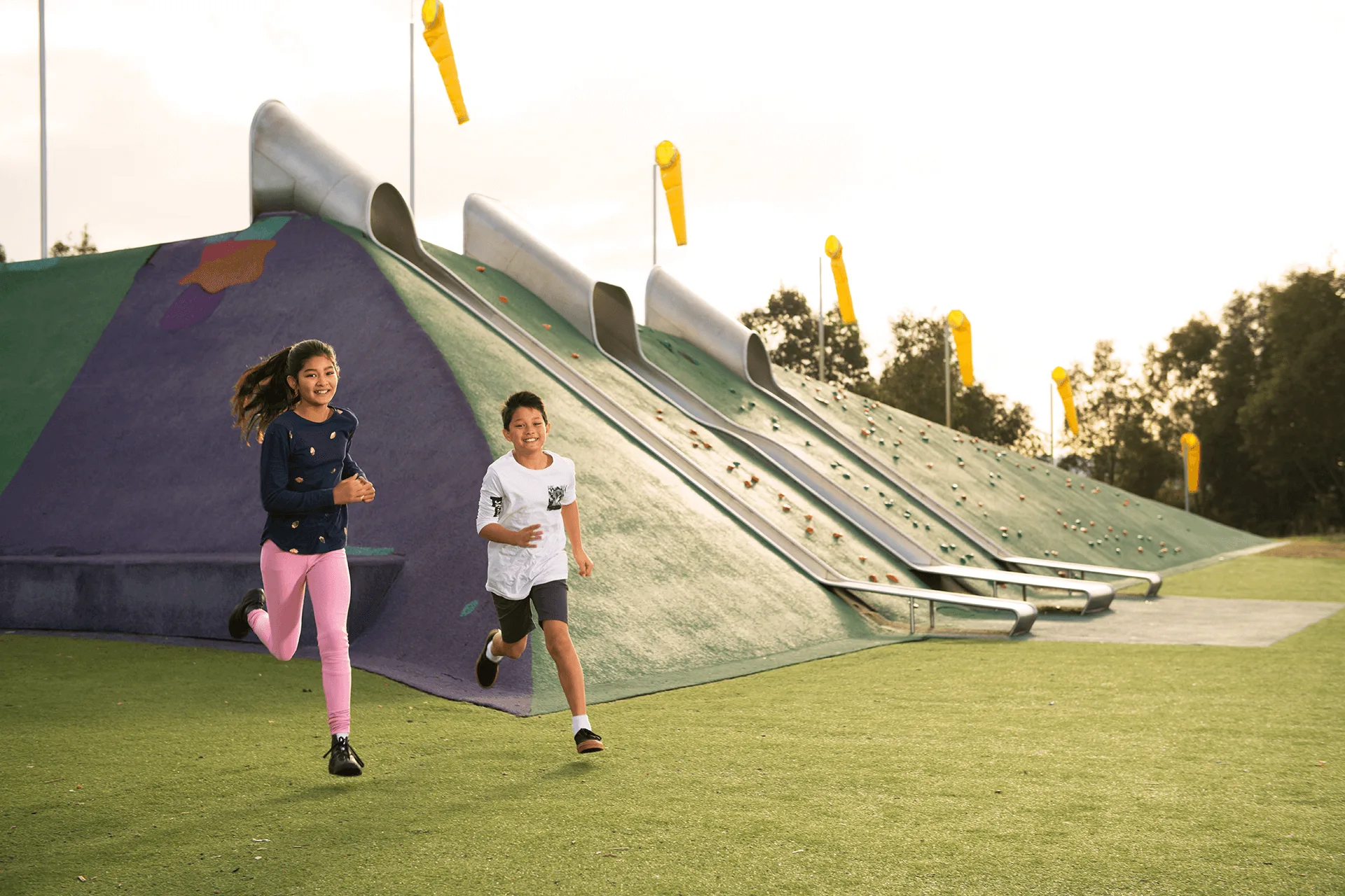 Two kids happily running on artificial turf next to a large play hill featuring long metal slides and climbing holds.